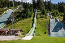 Skispringen in Ramsau am Dachstein - Ramsau am Dachstein ist Weltcup-Standort für die Skisprung-Damen und auch für die nordische Kombination der Herren. Die Schanzenanlage verfügt über Matten für den Sommer und auch ein Kuriosum, das es so wir wissen, nur einmal auf der Welt gibt: Eine Anlauf-Testspur, die hier in der Mitte zu sehen ist. • © alpintreff.de - Christian Schön